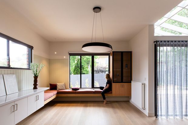Bright contemporary open-plan living and dining area with pale timber flooring, white built-in cabinetry, a window seat, and large glazed openings. A black oval pendant light hangs centrally, while sheer curtains and a roof-level skylight bring in abundant natural light. The room has warm neutral finishes and integrated timber-and-glass joinery.
