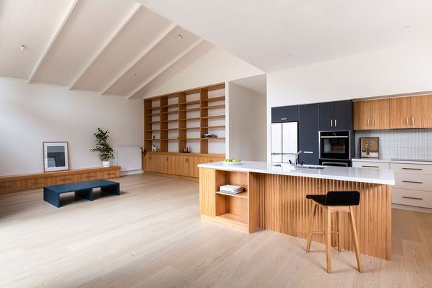 Bright open-plan contemporary kitchen and living area with pale timber flooring, a large fluted timber kitchen island, black and timber cabinetry, integrated appliances, open display shelving, and a built-in timber media/storage wall. The space has a high pitched ceiling with exposed painted beams and a restrained neutral palette.