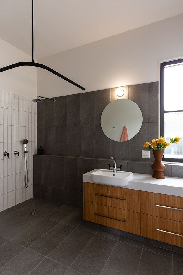 Contemporary bathroom with a walk-in shower, dark grey tiled walls and floor, a white tiled shower niche wall, and a floating timber vanity with white benchtop and integrated basin. A round wall mirror, black shower fittings, window, and sculptural pendant-style light complete the space.