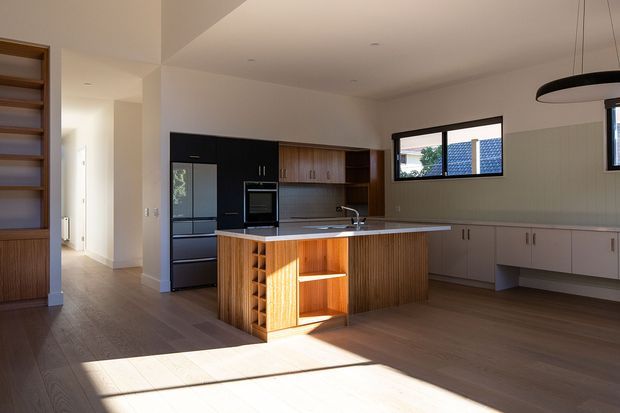 A spacious contemporary kitchen with an open-plan layout, timber flooring, and a central island featuring vertically grooved timber cabinetry, open shelving, and integrated wine storage. The kitchen includes dark cabinetry and appliances on one side, warm timber overhead joinery, pale lower cabinets, a light benchtop, a subtle vertical-panel splashback, and broad black-framed windows. A minimalist circular pendant light hangs above the island, while built-in shelving and a hallway are visible at the left.