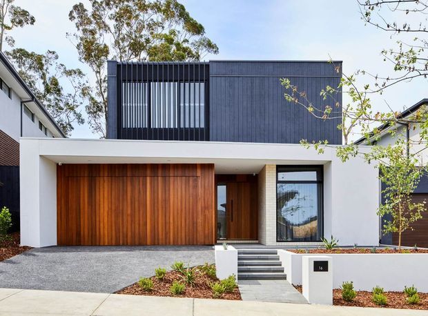 Street-facing exterior of a contemporary two-storey residence with a crisp white rendered facade, charcoal vertical cladding and screening, a broad flat roof, warm timber garage and entry doors, and a recessed entrance reached by concrete steps. The home sits in a suburban streetscape with restrained landscaped planting and a gravel driveway.