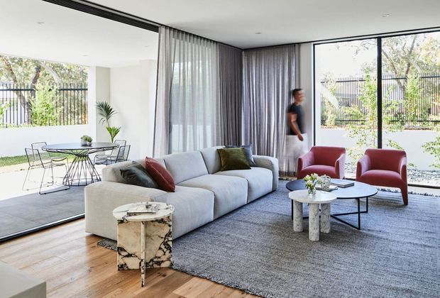 Bright, open-plan contemporary living room with floor-to-ceiling glazing, sheer curtains, a large pale sectional sofa, sculptural coffee and side tables, and two red lounge chairs. A dining area is visible behind the living space, with timber flooring transitioning to a large grey rug. The room opens visually to a landscaped exterior through expansive black-framed windows.