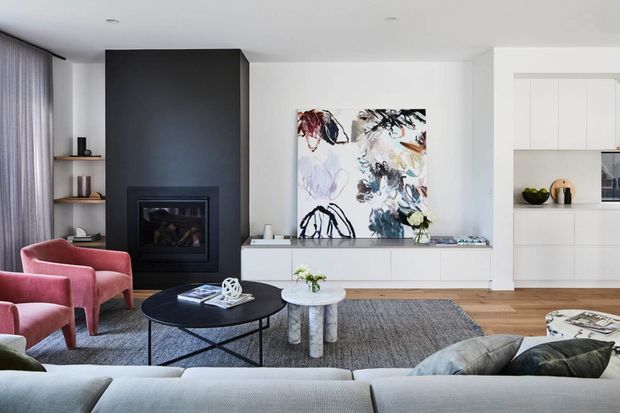 Bright contemporary living room with a large pale sectional sofa, pink upholstered armchairs, black round coffee table, textured rug, built-in white media cabinetry, and a bold abstract artwork. A dark feature wall incorporates a modern fireplace, while open timber shelving and an adjoining white kitchen are visible in the background.