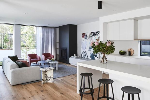 Bright open-plan contemporary living and kitchen area with pale timber flooring, white cabinetry and island, black stools and fireplace surround, large glazed doors, neutral furnishings, and a colourful botanical artwork feature wall.