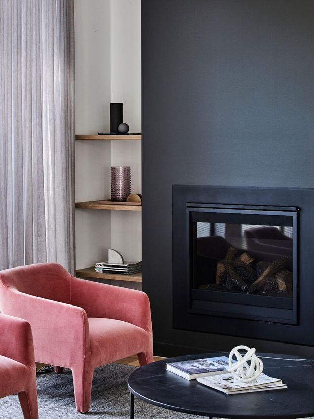A refined living room corner featuring a dark charcoal fireplace wall with a built-in gas fireplace, open timber display shelving, sheer floor-to-ceiling curtains, plush dusty-rose velvet armchairs, and a low round black coffee table with magazines and sculptural decor. The space has a calm, sophisticated contemporary aesthetic with layered textures and muted contrast.