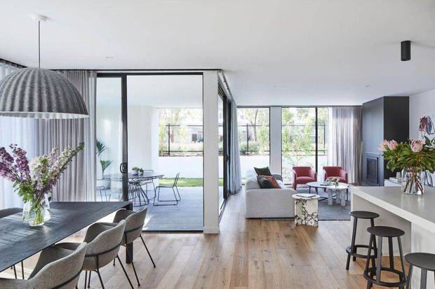 Bright open-plan contemporary interior combining a dining area, kitchen island and living room. The space features light timber flooring, white walls and cabinetry, extensive full-height glazing, sheer curtains, a sculptural pendant over the dining table, and colourful upholstered furniture and floral arrangements. A covered outdoor terrace and landscaped garden are visible beyond the glazing.