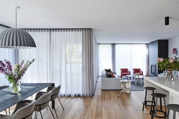 Bright open-plan contemporary interior combining a dining area, living room and kitchen. The space features light timber flooring, sheer full-height curtains, a dark dining table with upholstered chairs, a pale sofa, red accent lounge chairs, a minimalist kitchen island with stools, and a dark fireplace or media wall. Large glazed openings provide abundant natural light.
