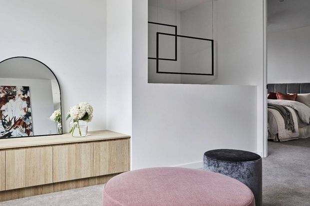 Contemporary bedroom-adjacent living area featuring a pale timber low sideboard, large arched mirror, abstract artwork, flowers, and sculptural upholstered ottomans. A partially visible bedroom with layered neutral bedding opens directly from the space, while simple black geometric wall art adds contrast to the light interior.