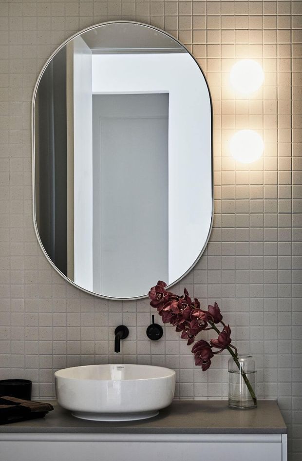 A refined bathroom vanity featuring a white vessel basin on a taupe-grey countertop, matte black wall-mounted tapware, and a large rounded-rectangle mirror. The wall is finished in small square light-grey tiles, with two spherical wall lights beside the mirror and a dark orchid arranged in a clear glass vase.