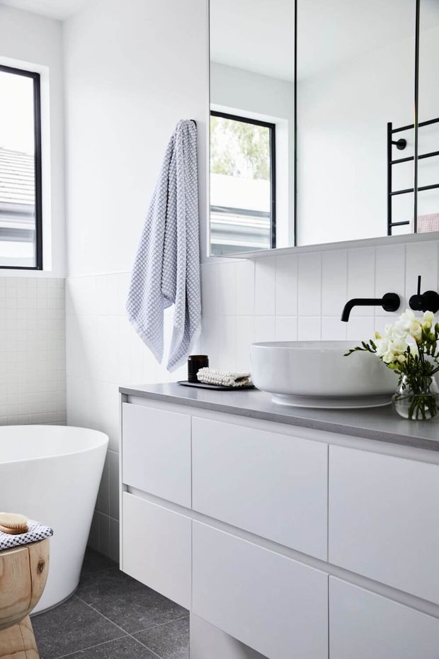 Bright contemporary bathroom featuring a freestanding white bathtub, wall-mounted white vanity cabinetry, grey countertop, white vessel basin, matte black tapware and heated towel rail, large mirror panels, white vertical wall tiles, and dark grey floor tiles.