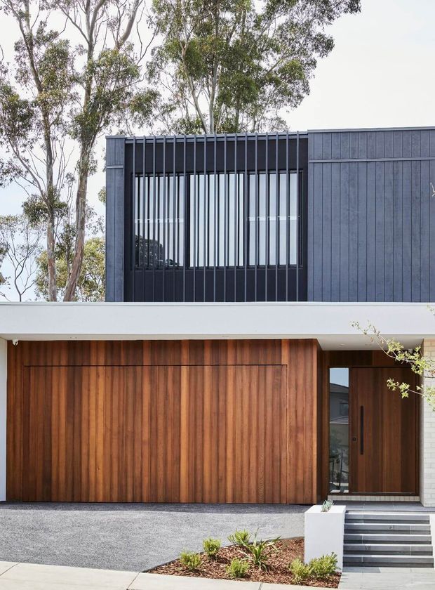 Contemporary two-storey residential facade featuring dark vertical cladding and a screened upper-level window, a broad white horizontal slab, and warm timber-clad garage and entry doors. The house is set against mature trees with a minimalist gravel driveway, concrete steps, and sparse landscaped planting.