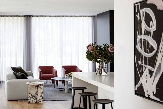 Bright contemporary open-plan living and kitchen area with floor-to-ceiling sheer curtains, a white sectional sofa, burgundy armchairs, marble coffee tables, and a large white kitchen island with dark stools. A bold monochrome abstract artwork provides a strong accent on the wall.