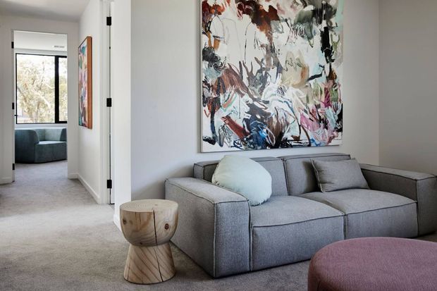 A bright, contemporary living room with a large grey sectional sofa, sculptural wooden side table, and mauve upholstered ottoman. An abstract artwork provides a colourful focal point above the sofa, while an open doorway reveals a carpeted adjoining room with additional seating and artwork.