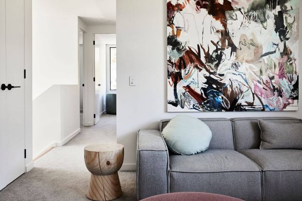 Bright contemporary living room with a large grey upholstered sofa, round pale blue cushion, abstract multicolour artwork, and a sculptural timber side table. A carpeted hallway and white interior doors extend behind the seating area.