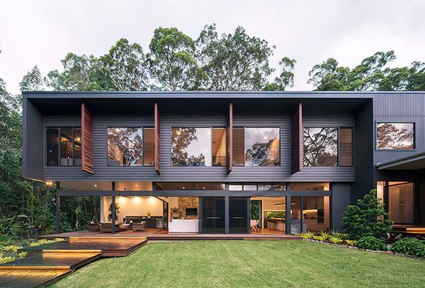 Contemporary two-storey dark-clad residence viewed from the landscaped rear garden. The facade features extensive glazing, projecting timber sun-shading screens, horizontal cladding, and a covered ground-floor entertaining area opening onto a timber deck and lawn.