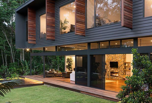 A contemporary multi-level home nestled in a lush wooded setting, featuring dark horizontal cladding, projecting timber-lined window boxes, extensive glazing, and a raised timber deck. The lower level opens to an outdoor living area, while the warmly lit interior includes a striking natural stone fireplace wall. Dense greenery surrounds the landscaped lawn and deck.