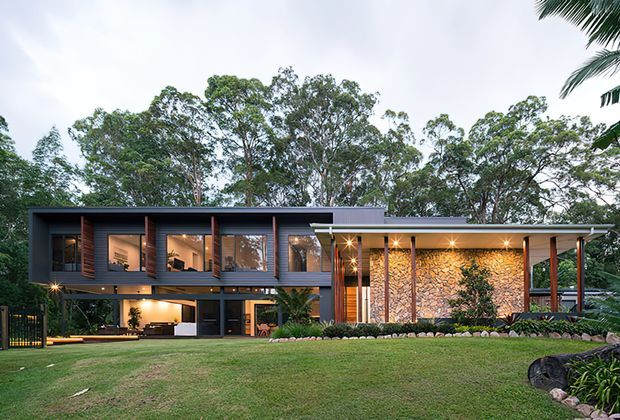 A contemporary two-storey residence set within a leafy landscape, featuring a long horizontal composition, dark cladding, extensive glazing, warm timber accents, and a prominent illuminated natural-stone feature wall beneath a broad roof overhang. The elevated upper level and open lower level create a lightweight modern form.