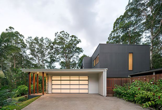 Exterior view of a contemporary multi-level home set within dense tropical greenery. The building features dark vertical cladding, a projecting upper volume, a low flat roof over the garage and entry, warm timber posts and screening, and a wide translucent-panel garage door. A concrete driveway and lush planted garden frame the facade.
