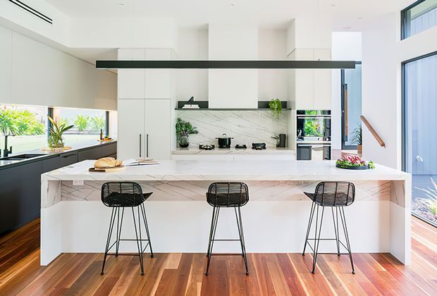 Bright contemporary kitchen with a large white marble-look island and three black wire-frame bar stools. White full-height cabinetry surrounds a cooking zone with a veined stone splashback, integrated appliances, indoor plants, and expansive glazing. Warm timber flooring contrasts with the predominantly white fit-out, while a stair and tall windows are visible at right.