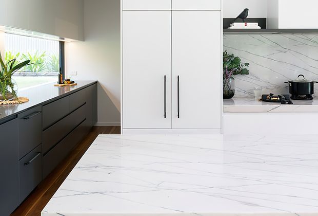 A contemporary kitchen featuring dark matte cabinetry along the window wall, tall white pantry cabinetry, and a large white island with subtle grey veining. The splashback uses matching white marble-look surfaces, with integrated appliances and minimal black hardware. Natural light enters through the window, creating a clean, refined atmosphere.