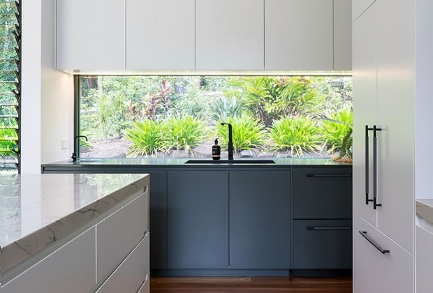 A bright contemporary kitchen with matte dark lower cabinetry, white upper cabinets, a large pale stone island benchtop, integrated appliances, and a wide picture window overlooking lush planting. Timber flooring and black hardware add contrast, while the generous glazing creates a strong indoor–outdoor connection.