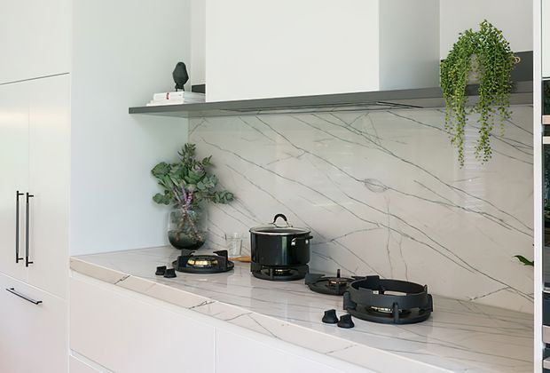 Bright contemporary kitchen with white flat-panel cabinetry, a large white marble-look stone benchtop and matching full-height splashback, a dark induction cooktop, and a slim dark rangehood shelf. Minimal decor and a trailing plant add subtle contrast.