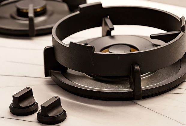 Close-up product-style view of a black gas cooktop burner and trivet assembly on a light countertop, with two separate black control knobs in the foreground and another burner partially visible in the background.