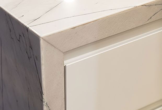 Close-up detail of a pale stone-look benchtop or countertop meeting a vertical side panel and light-coloured built-in cabinetry. The surface has subtle grey veining and a matte-to-honed appearance, with clean contemporary detailing.
