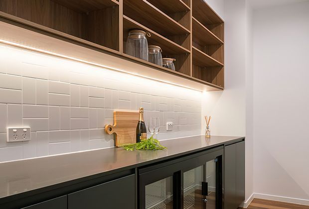 A refined kitchen or entertaining nook featuring dark built-in cabinetry with integrated glass-front beverage fridges, a grey stone benchtop, white rectangular tiled splashback, and warm timber open shelving illuminated by under-shelf lighting. Decorative glassware, bottles, and a small serving board sit on the counter, creating a polished bar-like preparation area.