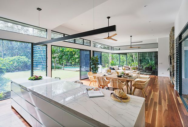 Bright, open-plan contemporary kitchen and dining area with extensive floor-to-ceiling glazing, a large white marble-look island, white cabinetry, warm timber flooring, and a timber dining table with woven chairs. Black linear pendant lights and ceiling fans contrast with the light interior, while lush greenery and a swimming pool are visible outside.