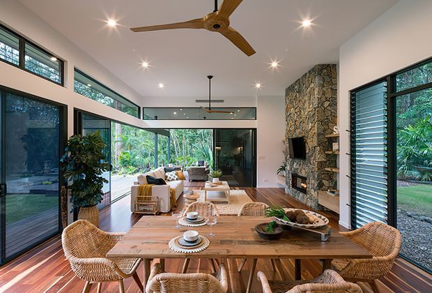 Bright open-plan living and dining space with floor-to-ceiling glazing, timber flooring, a large natural stone feature fireplace wall, and an open kitchen island in the foreground. Woven rattan dining chairs, timber furniture, indoor plants, and views to lush greenery create a warm, nature-connected contemporary interior. A high ceiling with recessed lighting and a large ceiling fan adds to the airy sense of space.