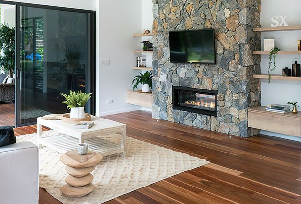 Bright contemporary living room with a substantial natural stone feature wall incorporating a television and linear gas fireplace. Built-in timber shelving and cabinetry flank the feature wall, while dark timber flooring, a large textured rug, light upholstered seating, a glass coffee table, indoor plants, and expansive dark-framed glazing complete the room.
