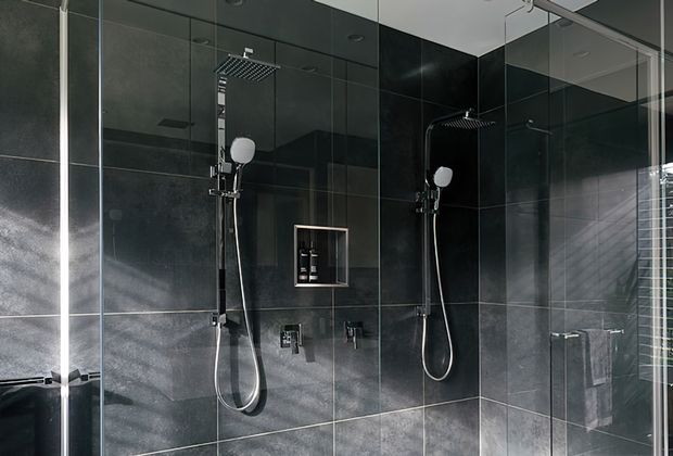 A modern bathroom shower enclosure with two separate shower stations, large square rainfall showerheads, handheld sprays, recessed wall niche, and dark glossy wall tiles. Frameless glass panels and reflective surfaces create a sleek, high-contrast finish.