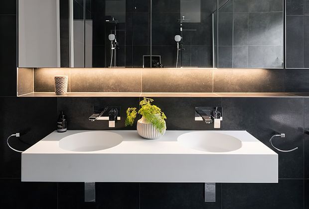Contemporary double-vanity bathroom with a floating white benchtop, two integrated oval basins, wall-mounted chrome tapware, dark large-format wall tiles, and mirrored cabinetry. A warm linear under-cabinet light creates contrast against the dark finishes, with a small decorative plant and accessories on the vanity.