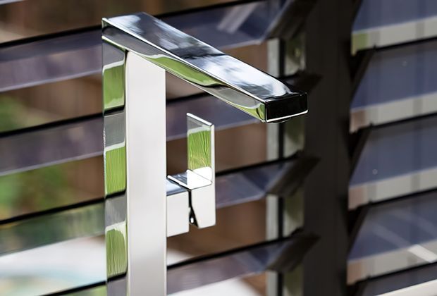 Close-up product photograph of a contemporary rectangular bathroom faucet with a chrome-silver polished finish and a matching single lever handle. The angular tap is mounted against a reflective horizontal slatted wall surface, with strong geometric lines and mirrored reflections.