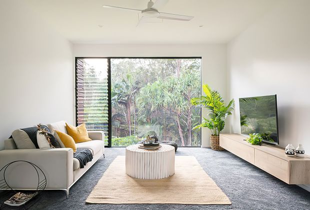 Bright contemporary living room with a large black-framed window overlooking lush vegetation, a neutral sofa with mustard and patterned cushions, a round fluted coffee table, pale rug, light timber media console, wall-mounted television, indoor plants, and a white ceiling fan.