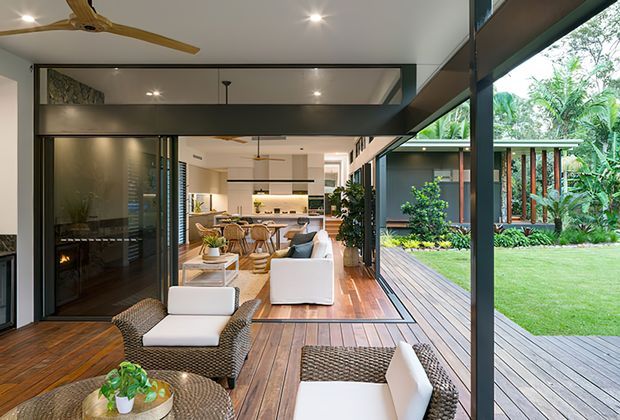 A spacious contemporary open-plan living area opening onto a timber deck and landscaped garden through extensive floor-to-ceiling glazing. The room includes woven lounge chairs, white upholstered seating, timber flooring, a dark fireplace wall, a dining area, and a kitchen visible in the background. Large structural steel or dark-framed glazing elements create a strong indoor-outdoor connection.