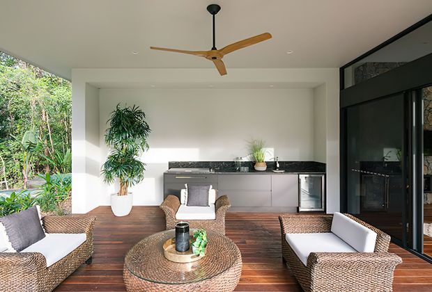 Bright open-plan indoor-outdoor living area with a timber deck floor, woven rattan seating and round coffee table, a compact contemporary kitchen with dark stone benchtop and grey cabinetry, extensive black-framed glazing, tropical planting, and a timber-bladed ceiling fan.