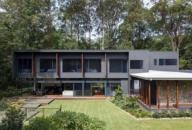 Contemporary multi-level home set within a lush bushland landscape. The building features a dark horizontal upper volume with extensive glazing, projecting timber-toned vertical fins, a lighter rendered side wing, and a lower outdoor living area opening onto a lawn and landscaped garden. A separate covered pavilion with stone walls and timber posts sits to the right.