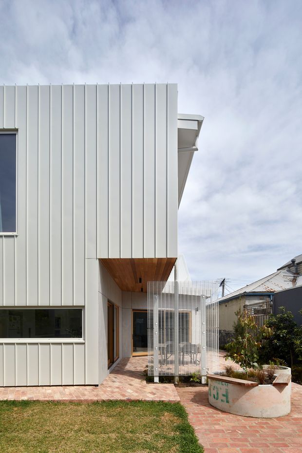 Exterior view of a contemporary multi-level residence with crisp white vertical metal cladding, a projecting timber-lined soffit, dark-framed glazing, and a brick-paved courtyard. A lightweight white screened outdoor structure and built-in curved planter add detail, with neighbouring homes visible beyond.