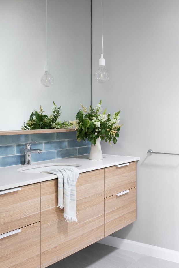 Bright contemporary bathroom vanity with a floating light-oak timber-look cabinet, white benchtop and integrated basin, chrome tapware, blue-grey rectangular tiled splashback, large mirror, and two clear globe pendant lights. A vase of greenery and a folded towel add softness to the otherwise clean, minimalist fit-out.