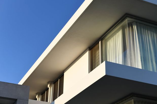 Close-up upward view of a contemporary white rendered building facade with strong horizontal slab lines, projecting balcony, floor-to-ceiling glazed openings, sheer curtains, and a flat roofline against a clear blue sky.