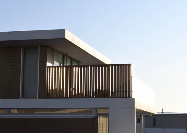 Exterior view of a modern two-storey residence with a flat roof, crisp white rendered volumes, dark horizontal cladding, extensive glazing, and a projecting upper-level balcony enclosed by vertical timber screening. A neighbouring building is partially visible in the background.