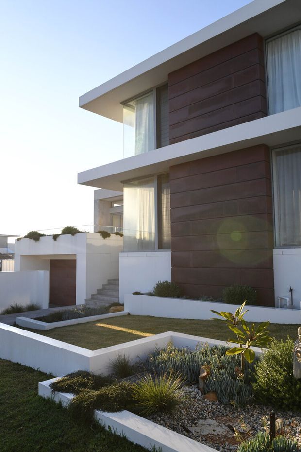 Exterior view of a modern multi-level residence with strong horizontal lines, projecting flat roof slabs, white rendered walls, large glazed openings with sheer curtains, and warm brown horizontal wall cladding. The front garden uses crisp white raised planter walls, gravel, lawn, and drought-tolerant ornamental planting.