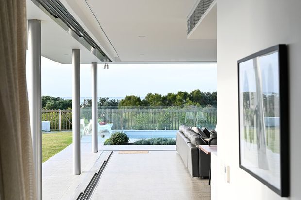 Bright contemporary open-plan living area opening directly onto a covered terrace and swimming pool. The space features white walls and ceiling, large retractable glazed openings, slender round columns, a pale tiled or concrete-look floor, and a dark sectional sofa. The exterior outlook includes landscaped greenery and a pool, while a framed artwork is visible on the interior wall.