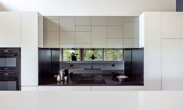 A sleek contemporary kitchen viewed straight on, with extensive white and pale grey flat-panel cabinetry, integrated black ovens at left, a dark benchtop and matching dark backsplash, undermount sink, cooktop, and a horizontal strip of windows above the work area. The restrained palette and handleless joinery create a minimalist, refined appearance.