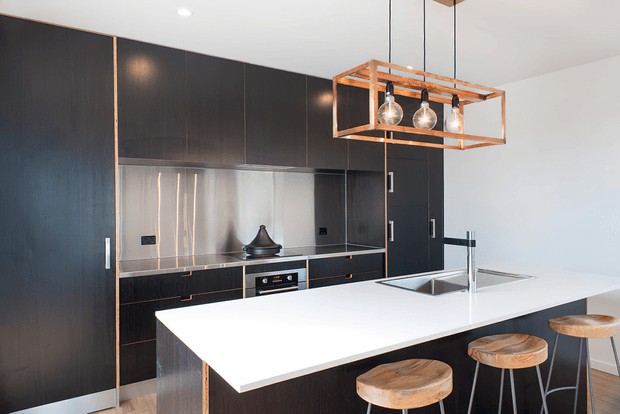 Contemporary kitchen with a large white island and integrated sink, dark full-height cabinetry, stainless-steel splashback, built-in appliances, and timber-topped stools. A copper-toned rectangular pendant light with exposed bulbs hangs above the island, creating a strong contrast against the monochrome cabinetry and white walls.