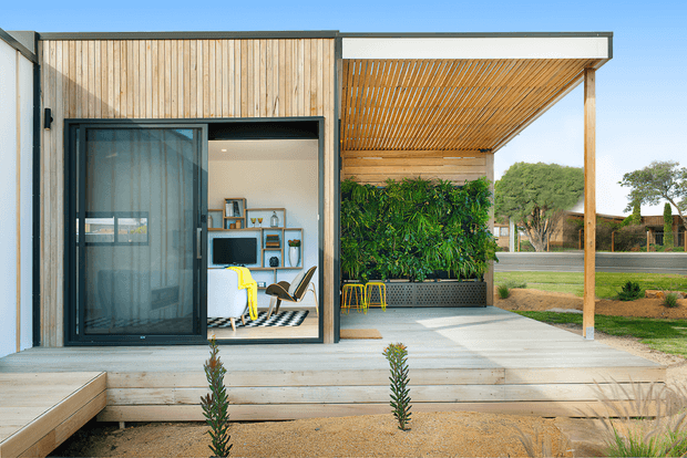 Contemporary single-storey pavilion-style exterior with a pale timber-clad facade, large black-framed glazed opening, and a covered outdoor terrace. The terrace features a slatted timber pergola, built-in raised planter with dense greenery, outdoor stools, and a light timber deck leading into a minimalist interior workspace or living area. The composition emphasizes indoor-outdoor connection and clean geometric forms.