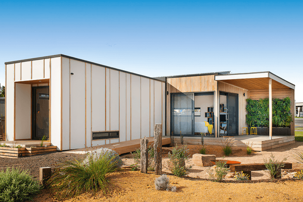 Single-storey contemporary modular-style building with clean rectangular forms, pale vertical panel cladding, warm timber-lined sections, broad glazing, and a covered recessed outdoor area. The building sits within a drought-tolerant landscaped garden featuring gravel, native grasses, low shrubs, timber edging, and stone elements.