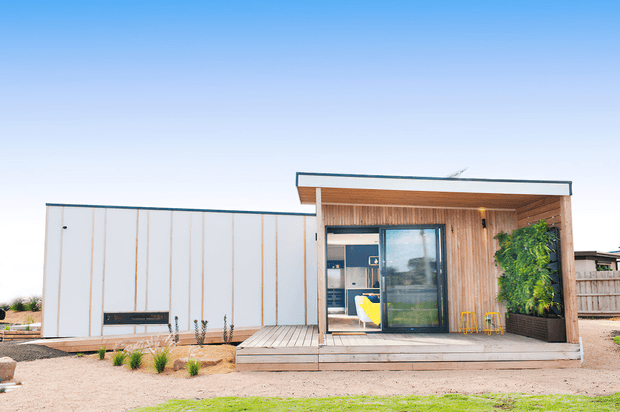Single-storey contemporary building with a crisp white-panelled volume adjoining a warm timber-clad entrance pavilion. The facade features a broad covered deck, large glazed sliding doors, vertical timber screening, integrated planter greenery, and a low flat roof, set within a sparse landscaped site.